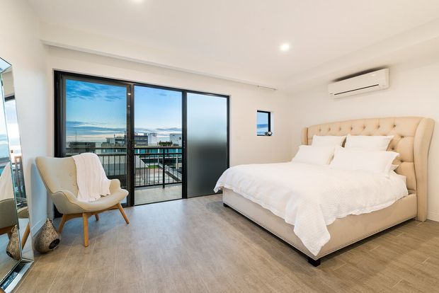 Bright contemporary bedroom with a large upholstered tufted bed, white bedding, pale timber-look flooring, recessed ceiling lighting, and a wall-mounted split-system air conditioner. Full-height black-framed glazing opens to a balcony and offers an expansive coastal or urban outlook, with a frosted sliding panel providing privacy. A light upholstered accent chair and full-height mirror sit near the glazing.