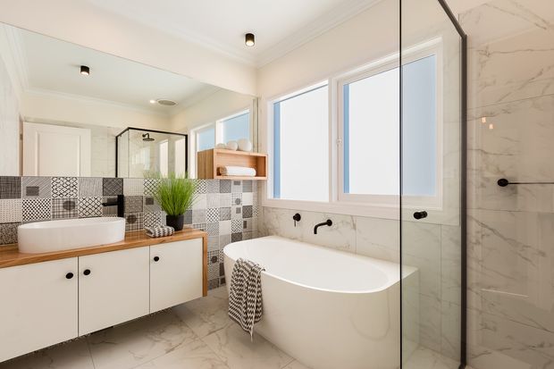 Bright, contemporary bathroom featuring a freestanding white bathtub beside a frameless glass shower, a floating white vanity with timber benchtop, patterned geometric wall tiles, marble-look floor and shower tiles, black tapware, and large frosted windows. A wall mirror expands the sense of space.