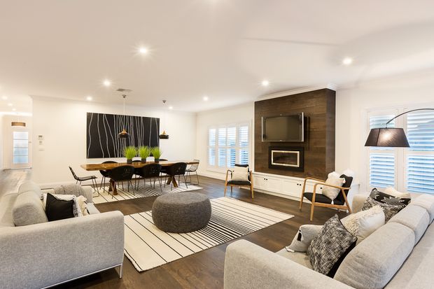 Bright open-plan living and dining area with a contemporary neutral interior. Two light grey sofas frame a striped rug and round upholstered ottoman, while a timber dining table with black chairs sits behind them. The room features dark timber flooring, white walls and ceiling, plantation shutters, recessed lighting, and a dark timber media wall with a television and built-in fireplace.