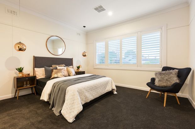 A bright, comfortably furnished bedroom with a low upholstered bed, layered neutral bedding, dark carpet, and a black lounge chair. White plantation shutters span the window wall, while warm pendant lights, a round wall mirror, and simple bedside furniture create a refined contemporary interior.