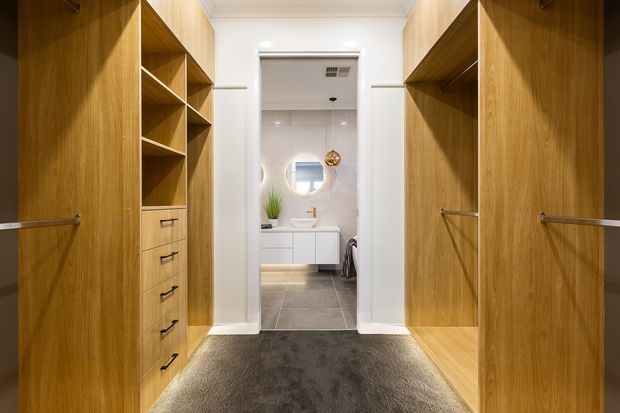A custom walk-in wardrobe with warm wood cabinetry lining both sides, open shelving, drawers, hanging rails, and a dark carpeted floor. A bright bathroom with a vanity, round mirror, tiled floor, and freestanding fixtures is visible through the doorway at the rear.