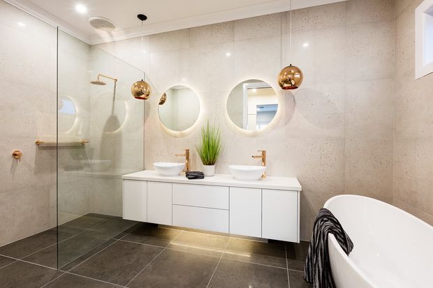 Contemporary bathroom with a spacious frameless glass walk-in shower, freestanding bathtub, floating white double vanity, round mirrors, and brushed brass fixtures. Large-format light stone-look wall tiles contrast with dark floor tiles, while warm pendant lights add a refined accent.