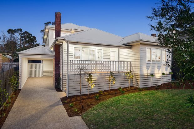 Exterior view of a single-storey suburban home with pale horizontal weatherboard cladding, a light grey pitched metal roof, prominent brick chimney, white louvred shutters and windows, a covered driveway leading to a garage, and a small landscaped front garden.