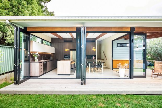 A single-storey contemporary pavilion-style extension with a broad low-pitched roof and fully openable glazed folding doors. The interior combines a compact dark timber kitchen with a central island, a bright dining area, white walls, exposed timber ceiling beams, and a timber deck opening onto a garden.