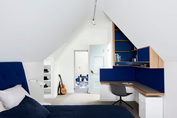 Bright attic bedroom with a sloped white ceiling, deep blue upholstered bedhead and bedding, and a custom built-in study nook. The study features navy-blue shelving and wall panels, warm timber work surfaces, white drawers, and a dark desk chair. Open shelving and a guitar are visible near the doorway, with a glimpse of a blue-and-white tiled bathroom beyond.
