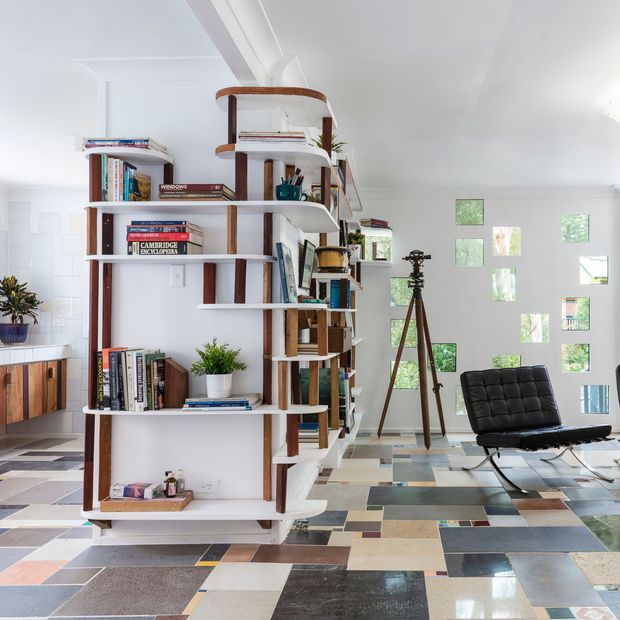 Bright open-plan contemporary interior featuring a sculptural freestanding white shelving unit with rounded shelves and dark timber supports, displaying books and plants. A black leather lounge chair and ottoman sit on a highly varied geometric patchwork tile floor. The room has white walls and ceiling, abundant natural light, a large patterned glazed wall, and a partial kitchen visible at left.