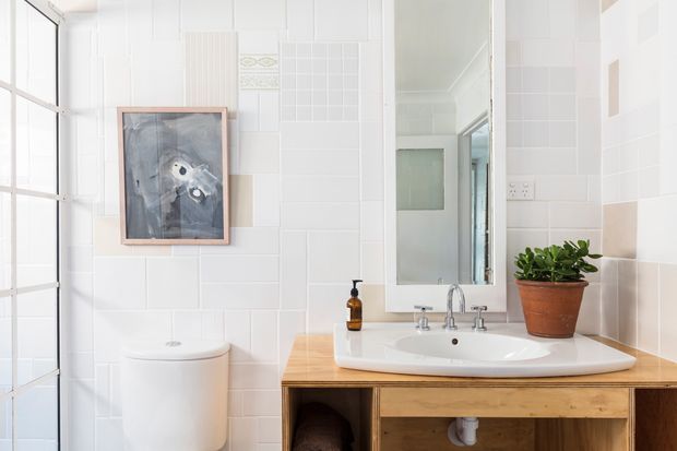 Bright, compact bathroom with white tiled walls, a wall-mounted mirror, white toilet and ceramic basin set into a warm timber vanity. A large grid-framed window or glazed door brings in abundant natural light, while a potted plant and framed artwork add restrained decorative detail.
