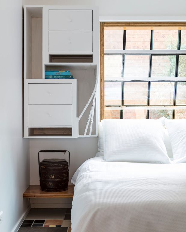 Bright, compact bedroom with crisp white bedding beside a large black-framed industrial-style window. A custom white built-in wall unit provides drawers, open shelving, and integrated storage, with a small timber bedside ledge and dark decorative container below. The floor features a geometric patterned tile finish, while exposed brick is visible beyond the window.