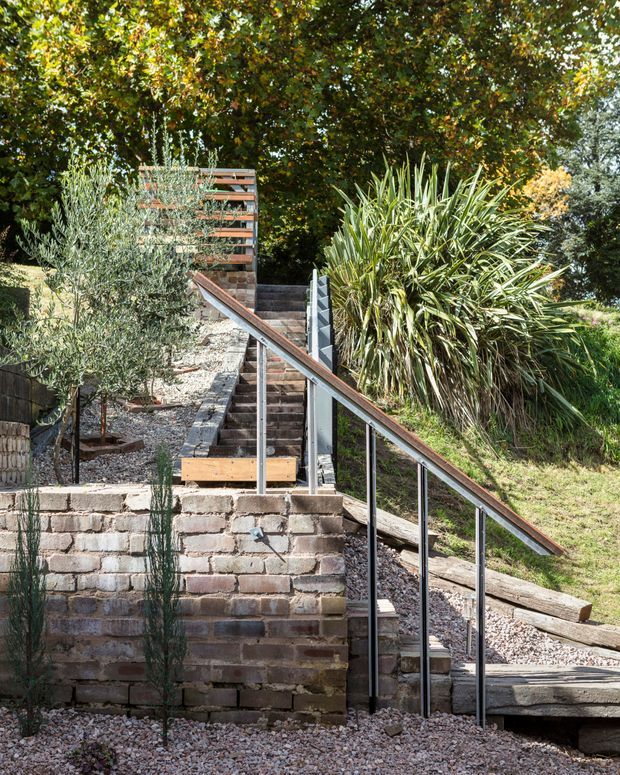 A landscaped sloped-site garden with a series of timber steps and terraces, supported by exposed aged brick retaining walls. A slim metal-and-glass balustrade follows the stairway, with gravel paths, timber edging, and dense mature planting including ornamental grasses, shrubs, and trees. The image focuses on the outdoor circulation and retaining landscape design.