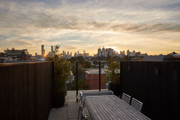 A rooftop or elevated outdoor terrace with dark timber privacy screens, a timber dining table and chairs, glass balustrading, and potted greenery. The terrace overlooks a dense urban skyline at sunset, creating a warm city outlook.