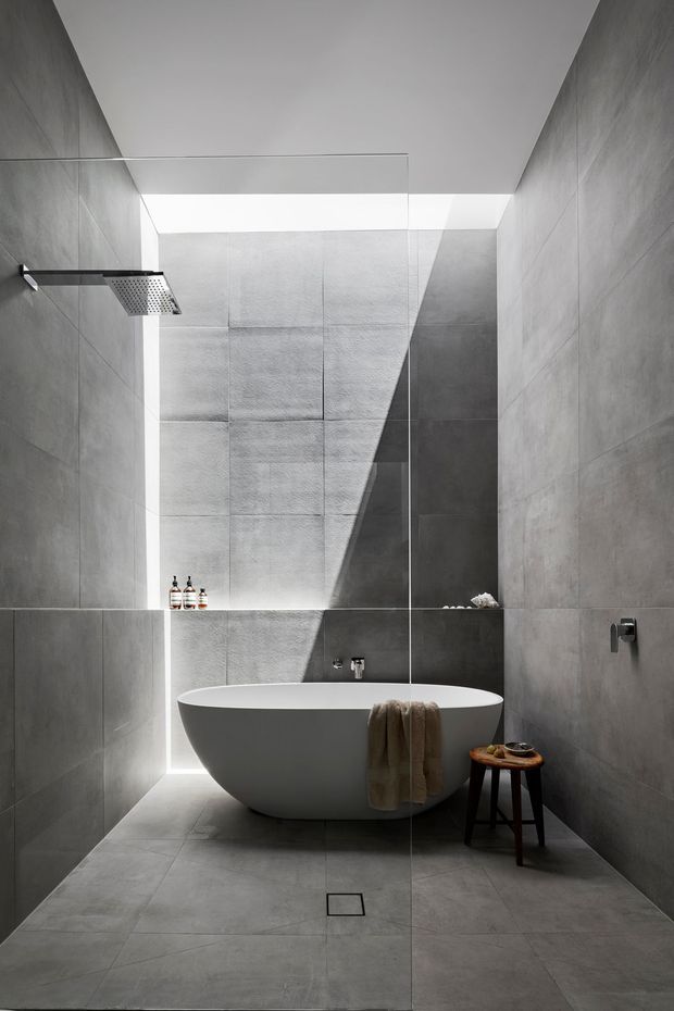 A refined contemporary bathroom featuring a freestanding white oval bathtub within a fully tiled grey enclosure. A frameless glass shower screen, wall-mounted rainfall shower, recessed shelf, and minimal fixtures create a clean architectural composition. Strong natural light enters from a high opening or skylight, casting a dramatic diagonal shadow across the rear wall. A small wooden stool and neutral towel add warmth to the otherwise monochromatic space.