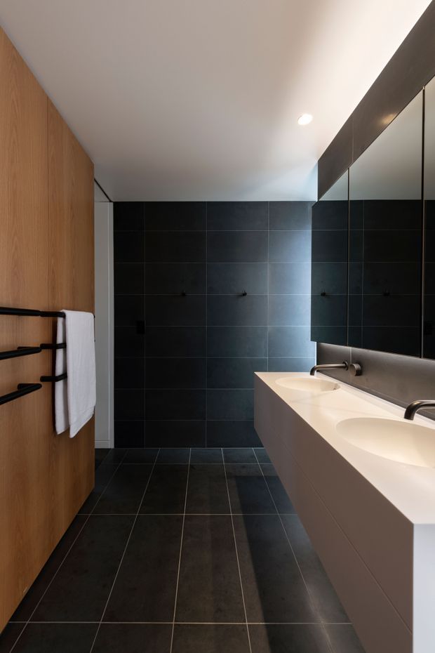 Contemporary bathroom with a long double vanity, integrated white basins and expansive mirrored cabinetry. Large charcoal-black tiled walls and floor contrast with warm timber wall panelling and black towel rails. Recessed ceiling lighting creates a clean, refined atmosphere.