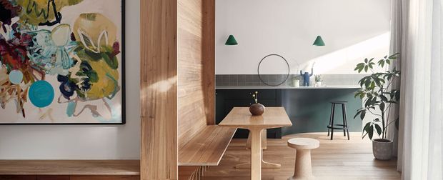 A bright, compact contemporary dining and kitchen area viewed through a timber-framed opening. The space features warm timber wall panelling and built-in bench seating, a small timber dining table, sculptural stool, dark kitchen cabinetry with a tiled splashback, pale flooring, green pendant lights, indoor plants, and abstract wall art. Sheer curtains and strong natural light create an airy, calm atmosphere.
