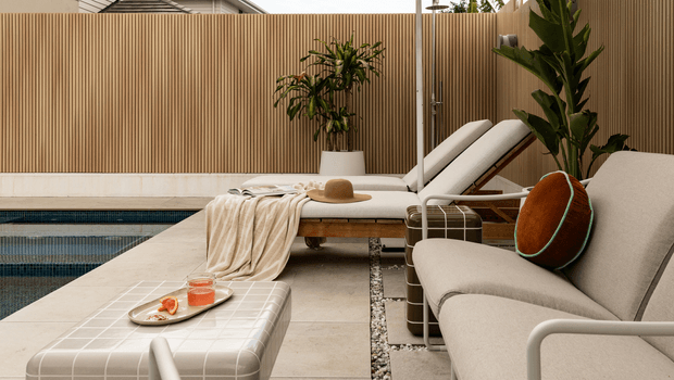 A refined outdoor poolside lounge area with a tiled swimming pool, pale stone paving, timber screening, reclining sun loungers, and a neutral upholstered sofa. A low rounded coffee table with a checked cover, refreshments, a throw, and cushions create a relaxed resort-like setting.