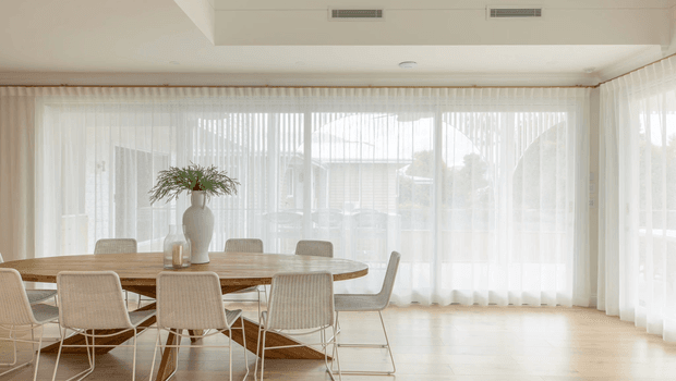 A bright, airy dining room with a large oval timber dining table surrounded by woven light-coloured chairs. Floor-to-ceiling sheer curtains span the windows and sliding glazing, while pale timber flooring, white walls, and a simple ceiling create a calm, minimalist interior. A large ceramic vase with greenery forms the table centrepiece, and the space opens toward a sunlit exterior through the glazing.