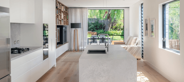 A bright, contemporary open-plan kitchen and living area. The kitchen features a large pale stone island with an integrated cooktop and sink, white flat-panel cabinetry, light timber flooring, and a stainless-steel refrigerator. A built-in dark fireplace or media feature is framed by timber shelving, while the rear living area has a light sofa and expansive glazing opening toward a leafy garden.