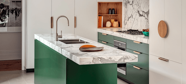 A refined contemporary kitchen centred on a large green island with a white marble-look benchtop, integrated sink and curved brushed-metal tap. Matching green cabinetry with slim brass-toned handles lines the rear wall, alongside a marble splashback, gas cooktop, built-in ovens and an open timber-lined display niche. Tall light neutral cabinetry, timber accents and pale tiled flooring create a warm, polished scheme.