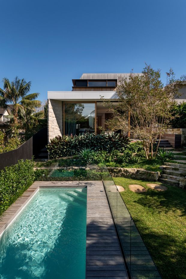 A landscaped residential exterior with a long narrow swimming pool in the foreground, bordered by dark timber decking and a glass balustrade. Stone garden steps, lawns, dense tropical planting and mature trees lead toward a contemporary multi-level house. The house features pale stone cladding, broad glazing, projecting flat roof forms, dark timber or metal details, and a vertically ribbed upper-level facade. Clear blue sky and strong sunlight create a warm, resort-like setting.