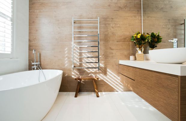 Bright contemporary bathroom with a freestanding white bathtub, wall-mounted timber vanity, white vessel basin, large mirror, chrome heated towel rail, and warm wood-look wall tiles. Strong sunlight enters through the window blinds, creating striped shadows across the room.