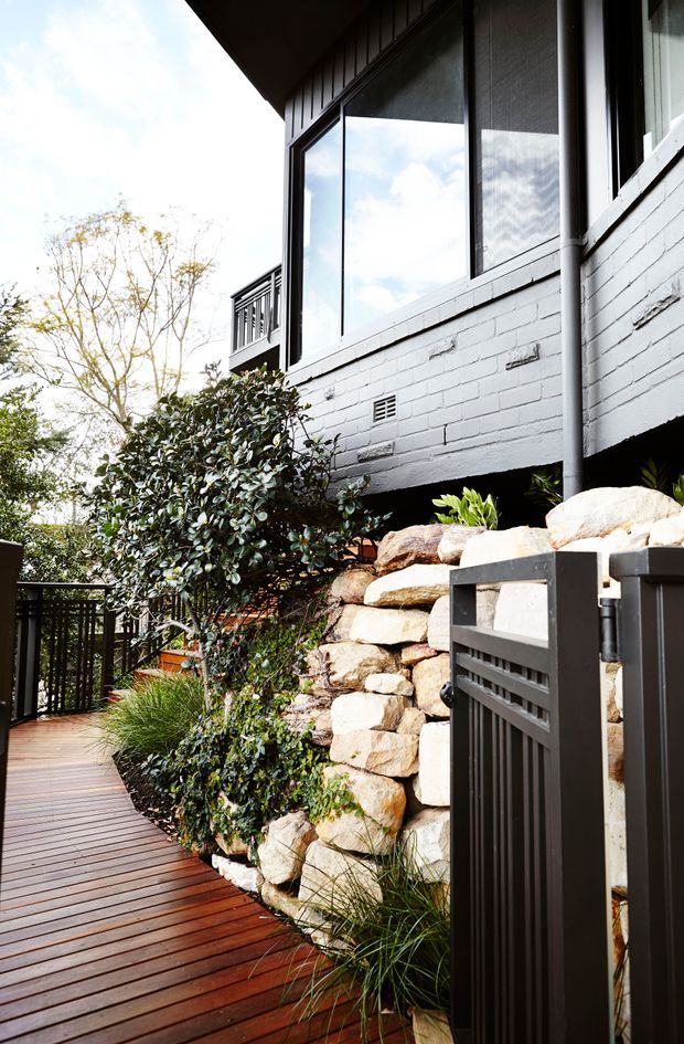A landscaped exterior walkway beside a raised house, featuring dark timber decking, a black metal gate and railing, a white-painted brick wall with large windows, and a natural stacked-stone retaining wall surrounded by dense planting.