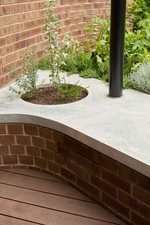 A landscaped courtyard or terrace detail with a curved raised brick planter wall, smooth pale concrete paving, integrated circular planting beds, dense greenery, and a dark cylindrical structural post. Timber decking occupies the lower foreground. The design combines robust masonry, concrete, planting, and curved forms in a contemporary outdoor setting.