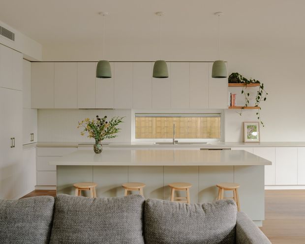 Bright open-plan kitchen viewed from behind a grey sofa. The room features a large white island with four timber stools, extensive white flat-panel cabinetry, a pale benchtop, a horizontal window above the sink, three green pendant lights, open shelving with plants and books, and light timber flooring. A living area is visible in the foreground.