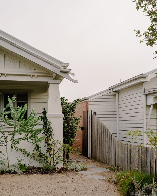 A narrow landscaped side passage between two single-storey residential buildings. The left cottage-style facade has pale horizontal weatherboards, a gabled roof with decorative eaves and a small window, while the right building has matching pale weatherboard cladding and a low-pitched roof. A timber paling fence, brick wall, gravel ground and stepping stones lead through the planted walkway. The scene has a soft, muted, residential character.