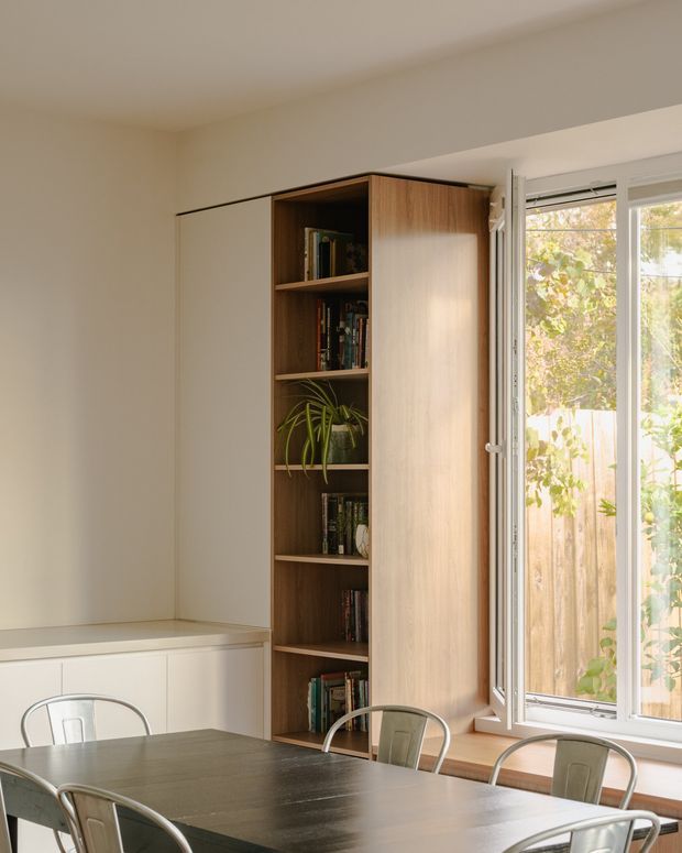 A warm, minimalist dining area with a long dark dining table and metal chairs in front of a built-in timber bookcase. The bookcase includes open shelving, books, and a potted plant, while a large white-framed window brings in natural light and overlooks a fenced garden. White walls, integrated cabinetry, and clean lines create a simple contemporary interior.