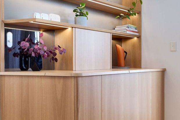 A close-up of a built-in timber storage and display unit, featuring light wood cabinetry, open shelving, integrated lighting, books, and decorative plants. The joinery has a clean contemporary appearance with a warm natural palette.