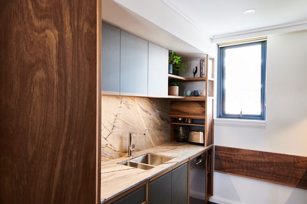 Compact contemporary kitchen or kitchenette with dark timber wall panelling, muted grey-blue cabinetry, and a dramatic veined stone benchtop and splashback. The space includes an undermount double sink, open timber shelving with small appliances and decor, integrated lower cabinetry, and a window providing natural light. White walls and ceiling create a bright, refined contrast with the darker wood finishes.