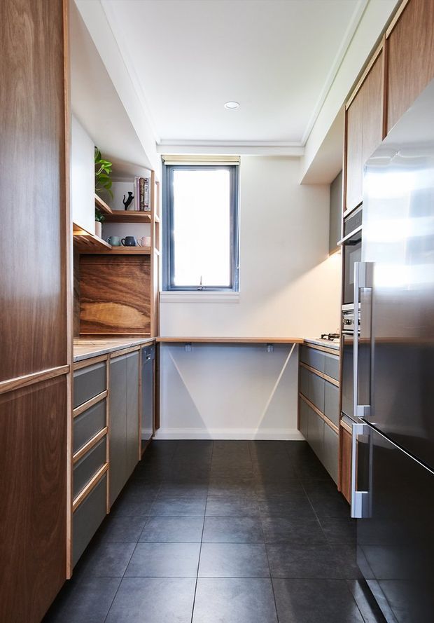 Compact galley kitchen with dark tiled flooring, warm timber cabinetry and open shelving, grey lower cabinets, stainless-steel appliances, and a bright central window. The room has a clean, contemporary built-in layout with a recessed ceiling detail and integrated refrigeration and ovens.
