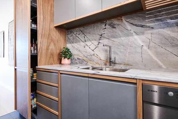 Contemporary galley-style kitchen with dark grey flat-panel cabinetry, warm timber accents, a large veined stone splashback and benchtop, stainless steel double sink and tap, integrated storage, and a built-in dishwasher. A narrow pantry with open shelving is visible at the left.