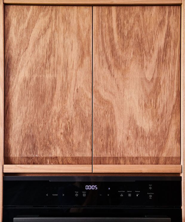 Close-up of a contemporary built-in kitchen appliance with warm natural timber cabinetry above a sleek black glass electric cooktop or oven control panel. The cabinet fronts feature prominent vertical wood grain and framed detailing, with subtle integrated controls and a digital display.