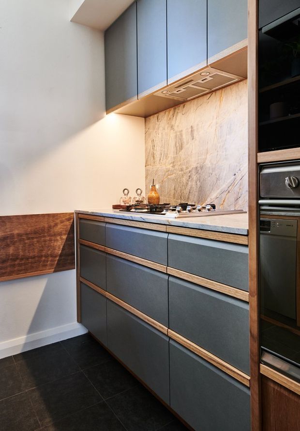 A compact contemporary kitchen with dark blue-grey flat-panel cabinetry accented by timber drawer trims and side framing. The kitchen features a pale veined stone splashback and benchtop, a gas cooktop with integrated rangehood, dark floor tiles, timber wall panelling, and built-in stainless steel ovens.