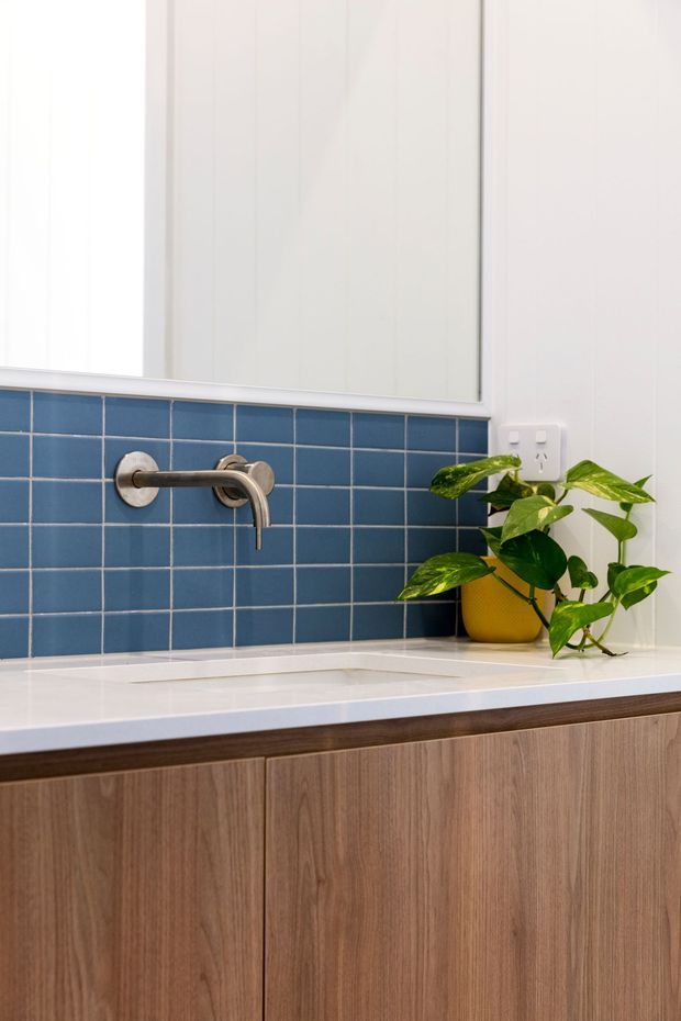 Close-up view of a contemporary bathroom vanity with warm wood-grain cabinetry, a white tiled benchtop, blue square-tile splashback, wall-mounted brushed metal tapware, and a white mirrored cabinet above. A potted trailing plant adds greenery beside the basin.