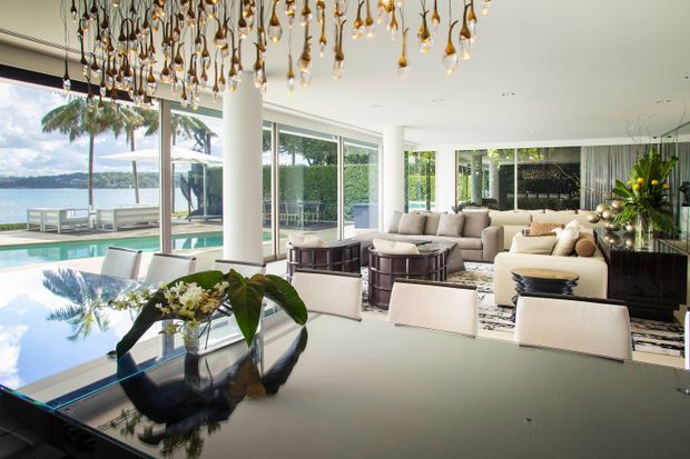 A bright, expansive contemporary open-plan living and dining area with floor-to-ceiling glazing overlooking a pool and waterfront landscape. The room features a glossy dark dining table, upholstered dining chairs, neutral sofas and armchairs, dark built-in cabinetry, and a dramatic sculptural pendant installation with numerous hanging glass-like forms. White structural columns, pale flooring, and abundant natural light create a luxurious indoor-outdoor atmosphere.