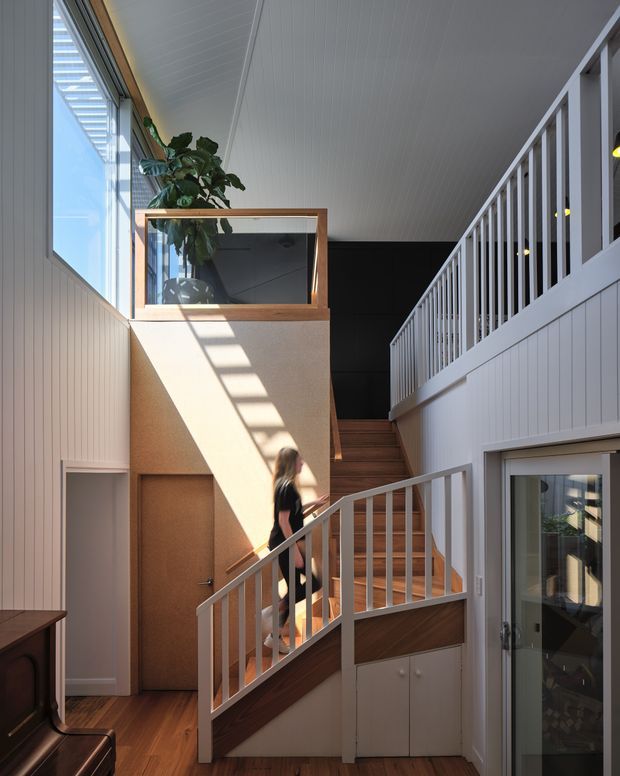 A bright, double-height residential stairway and landing with warm timber stairs and flooring, white vertical wall panelling, white balustrades, and a black feature wall at the upper level. Strong sunlight enters through high glazing, casting geometric shadows across the stair enclosure. A glazed timber display box with a plant sits above a doorway, and built-in cabinetry is visible beneath the stairs.