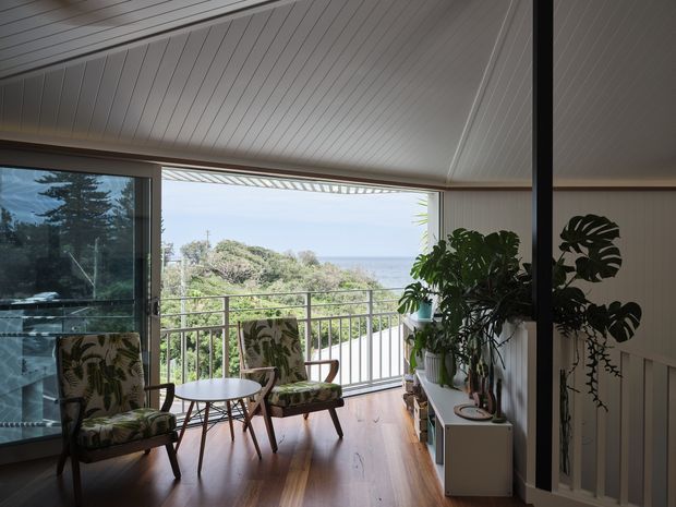 A bright, elevated sunroom or enclosed balcony overlooking lush vegetation and the ocean. The room features a sloped white panelled ceiling, warm timber flooring, extensive glazing, a metal balustrade, patterned upholstered lounge chairs, a small round table, and abundant indoor plants. A dark vertical structural post and stair or railing detail frame the right side.