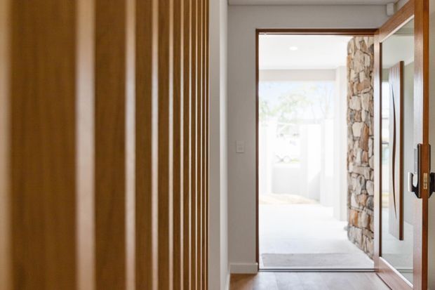 A bright residential hallway viewed toward an open doorway, with warm timber vertical screening on the left, timber-framed glazing or a mirrored panel on the right, a pale smooth wall and ceiling, and a prominent natural irregular-stone feature wall beyond the doorway. The corridor has timber flooring and a calm, modern natural-material palette.