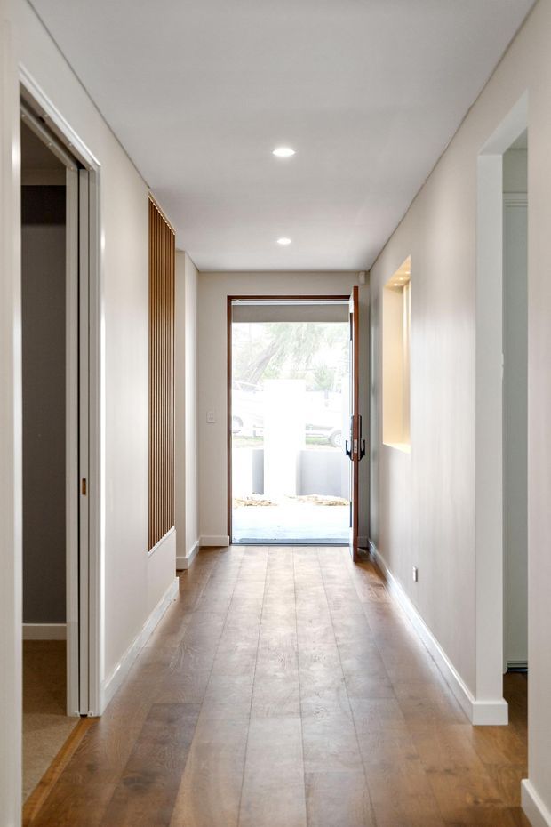 Bright, narrow residential hallway leading to a glazed front entry door. The corridor has pale painted walls, recessed ceiling lights, warm timber flooring, white skirting and trims, a vertical timber slat feature on the left wall, and doorways opening to adjacent rooms.