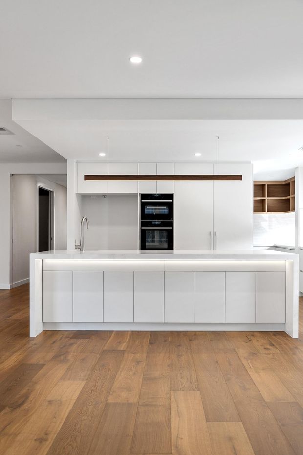 Bright contemporary kitchen viewed straight on, featuring a large white island with integrated cabinetry, white full-height wall cabinetry, a built-in black oven stack, stainless-steel sink tap, warm timber accent shelving and trim, and wide-plank timber flooring. The open-plan space has a clean white palette and recessed lighting.