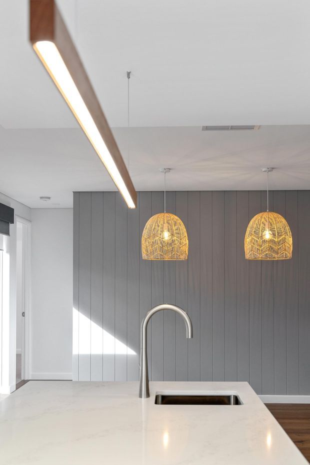 Bright contemporary kitchen featuring a large pale stone island with integrated sink and tall curved tap, a dark grey vertically panelled feature wall, warm woven pendant lights, and a linear timber-and-light fitting above the island. White walls, timber flooring, and strong daylight create a refined, modern interior.