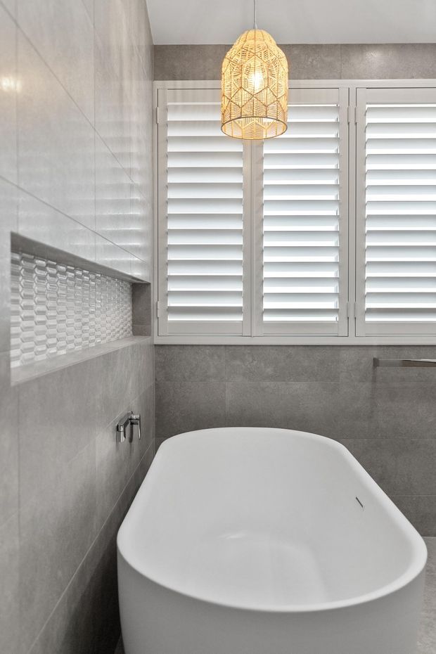 A contemporary bathroom featuring a freestanding white bathtub beneath a large shuttered window. Grey tiled walls and a recessed tiled niche with small textured mosaic tiles create a restrained neutral backdrop, while a woven pendant light adds warmth above the bath.