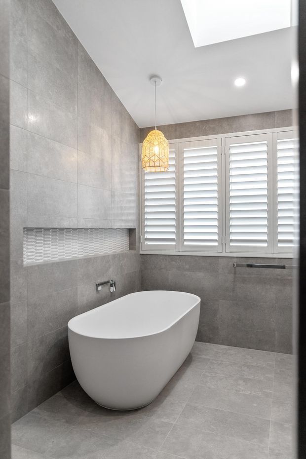 Modern bathroom featuring large-format grey stone-look wall and floor tiles, a freestanding white oval bathtub, a recessed mosaic-tile niche, white plantation shutters, and a warm woven pendant light.