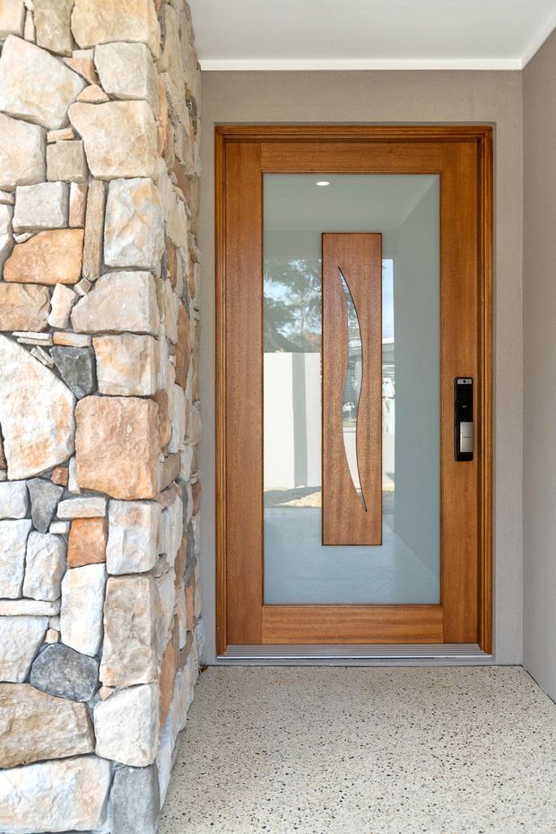 Close-up view of a contemporary residential entryway featuring a timber-framed glazed front door with a sculptural timber inset, dark electronic lock, and polished aggregate concrete floor. The entrance is bordered by a prominent irregular natural stone-clad wall and a smooth taupe painted wall.