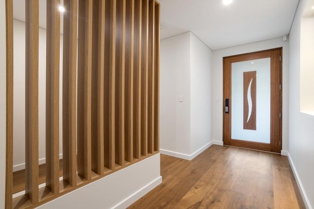 A bright residential entryway with warm timber flooring and a contemporary timber-and-glass front door. A full-height vertical timber batten screen creates a partial divider on the left, while white walls and ceiling provide a clean, minimalist backdrop.