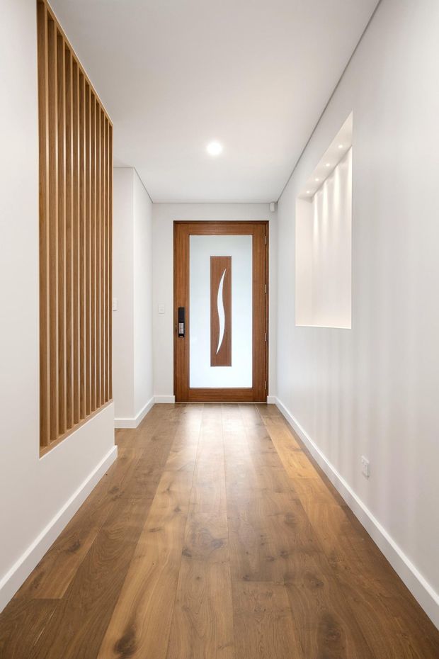 A bright, minimalist residential hallway with warm timber flooring, white walls and ceiling, a vertical timber slat screen on the left, recessed display niches with downlights on the right, and a timber-framed glazed entry door at the far end.