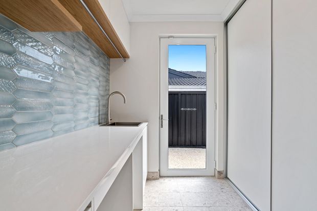 Compact modern utility or laundry space with white built-in cabinetry, a white benchtop, stainless-steel sink and tap, pale textured blue-grey tiled splashback, warm timber overhead shelving, and light stone-look flooring. A glazed external door opens to a private paved area.