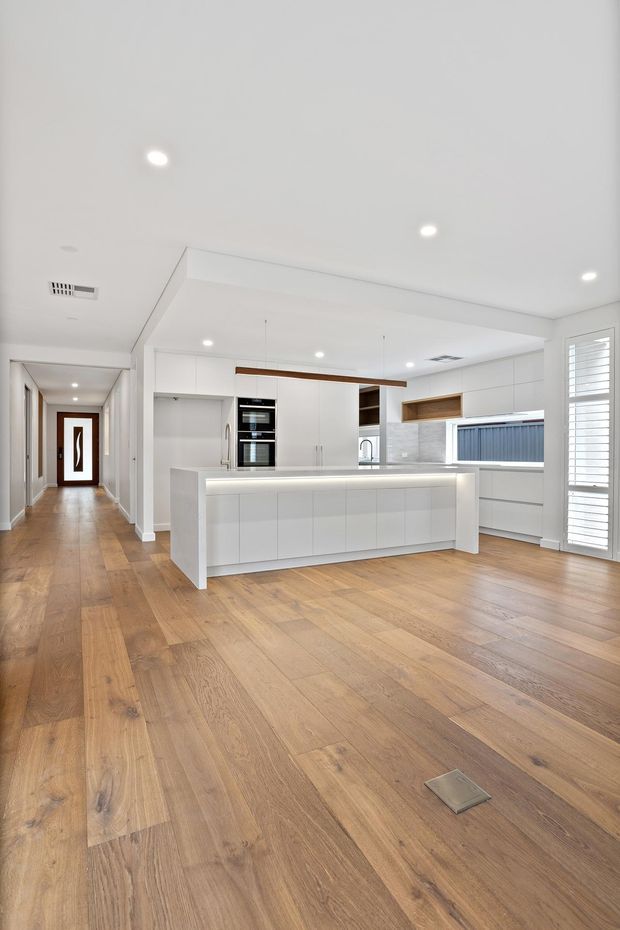 Bright open-plan kitchen viewed from a connected hallway, featuring white flat-panel cabinetry, a large central island with integrated lighting, white benchtops and splashback, timber floating shelves, built-in appliances, plantation shutters, and warm timber flooring. The space has a clean contemporary appearance with recessed ceiling lights and a simple linear pendant over the island.