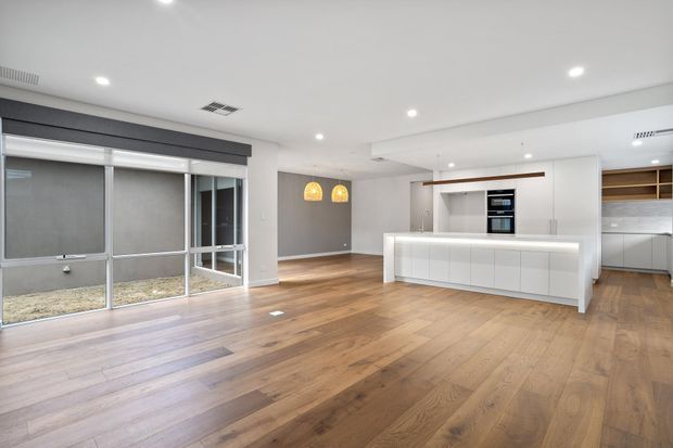 Bright open-plan contemporary interior featuring a large white kitchen island with integrated cabinetry and appliances, warm timber flooring, recessed ceiling lighting, expansive sliding glass doors, and a small dining area with pendant lights.